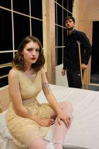 a theatre student sitting on a bed, another theatre student stands with a wooden crutch