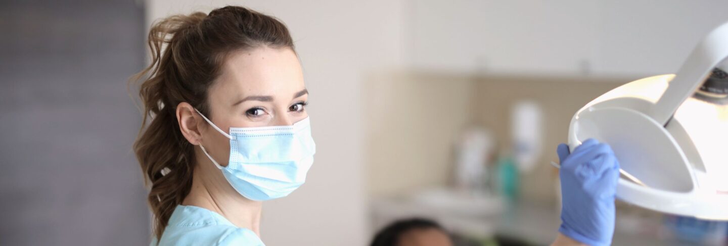 a woman wearing medical mask smiles at camera and adjusts dental light