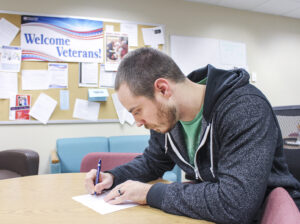 a student writes at a table with a sign reading veterans center in the background