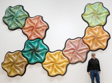 sculpture artist Chip Clawson posing in front his large artwork, taking up the space of an entire wall behind him